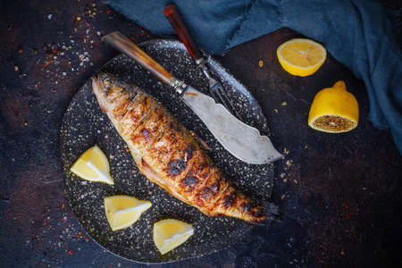 Top view of Grilled whole trout served on plate with lemon with seasoning on background. Knife near dish. Scattered spices. Dark mood photoの写真素材