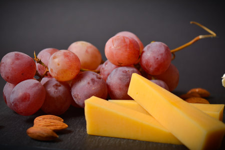 Side view of French hard cheese cheeseboard with pink grape and almond nuts on black slate stone plate. Dish served on black background. Close up of appetizers, startersの写真素材