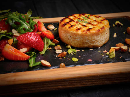 Grilled Camembert cheese steak served with almond and pistachio nuts. Fresh strawberry salad with arugula leaves near melted hot cheese. Whole Baked Cheese wheel on board on black wooden backgroundの写真素材