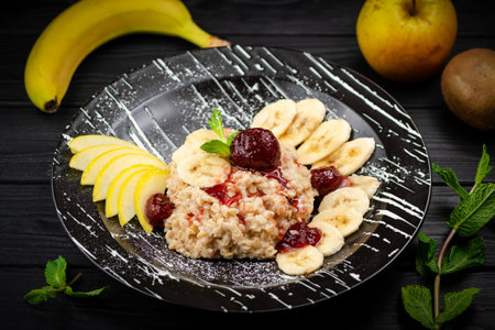 Healthy breakfast of Oatmeal porridge with banana, apple slices served with strawberry jam and fresh mint leaves. Dish on black wooden background with fruits out of focus.の写真素材