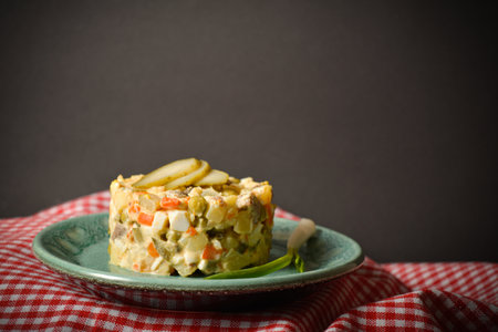 Side view of traditional rustic salad Olivier served in culinary rings Country Salad with potato, carrot, green peas, pickled cucumbers, eggs on American red checkered kitchen towel, black backgroundの写真素材