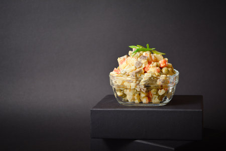 Close up to Traditional Russian salad Olivier served in glass bowl located on box. Salad with potato and carrot cubes, green peas, pickled cucumbers, eggs on Dark backgroundの写真素材