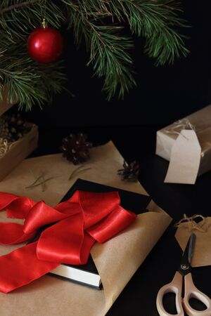 Christmas gift wrapping process. The book is packaged as a gift in craft paper with a wide red ribbon. Fir branch decorated with shiny colorful balls on the background. Happy new year themeの写真素材