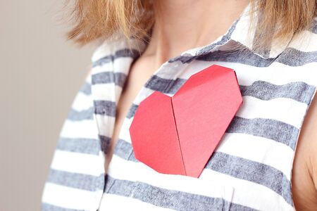 caucasian woman holding origami red paper heart, puts valentine in breast pocket. Insurance, charity donation concept. Place for text, copy space, selective focusの写真素材