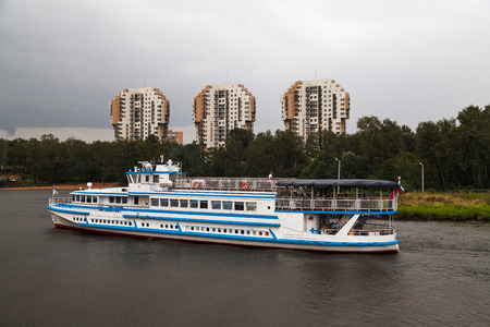 Cruise ship sets sail on the river by high-rise buildings and treesの写真素材