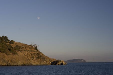 Full moon (daytime), rising over a shoreline with deep blue water in the foreground.  In the...