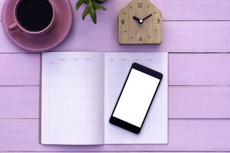 Mock up image of weekly planner, mobile smart phone with blank white screen ,home shaped clock and cup of coffee on pink wooden table background. Business technology concept.の写真素材