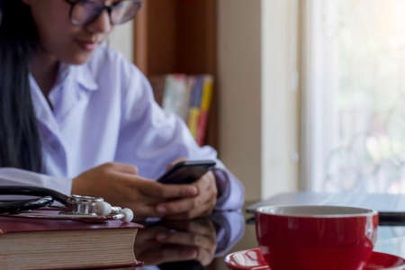 Female doctor in white coat, hand holding and using mobile smart phone with laptop computer, medical stethoscope and book on the desk at office. Medical online networking, telehealth concept.の写真素材