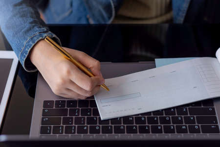 Businesswoman hand writing and signing blank checkbook and working on laptop, mobile phone on the wooden table at office. Business payment by cheque concept.の写真素材