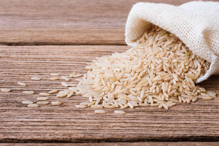 Coarse brown jasmine rice ( milled rice imperfectly cleaned, unpolished or half milled rice ) in hemp sack bag  isolated on old rustic wood table background. Healthy food and diet concept. Top viewの写真素材