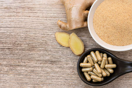 Dry ginger powder with capsules in  ceramic bowl and fresh ginger root with slice isolated on wood...