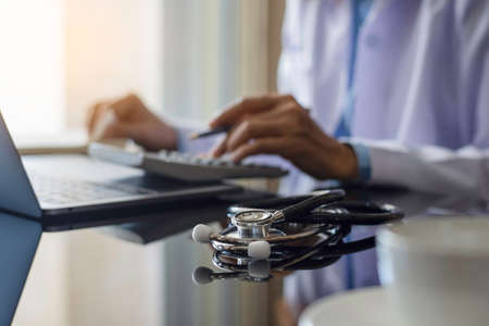 Doctor in white coat hand using calculator for account about medical costs with stethoscope and...