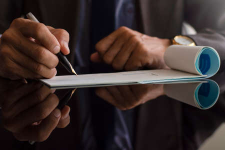 Business man in suit, hand write and signs checkbook on the dark mirror  desk at modern office. Paycheck and payroll concept. Payment by cheque. Copyspace with reflection.の写真素材