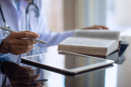 Doctor in white lab coat reading book and work on digital tablet  computer on the desk at office in medical room at clinic or hospital. Online medical learning concept.の写真素材