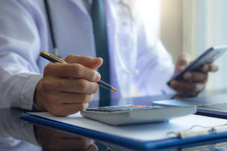 Male doctor hand hold pen, using calculator for calculate about medical costs and using mobile phone...