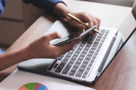Closeup hand work on laptop computer, use mobile smart phone with business graph data paperwork on...