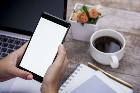 Mockup image of woman hand holding and using black mobile smart phone with blank white screen, work on laptop computer with tablet and cup of coffee on wooden table at home .の写真素材