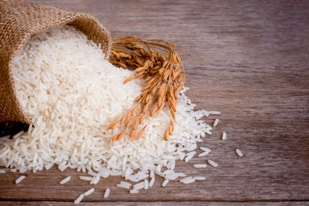 White rice in hemp sack bag with dry ear paddy pouring on old rustic wood table background. Healthy food and diet concept.の写真素材