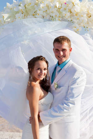 Happy smiling bride and groom in white embrace in front of the arch of flowersの写真素材