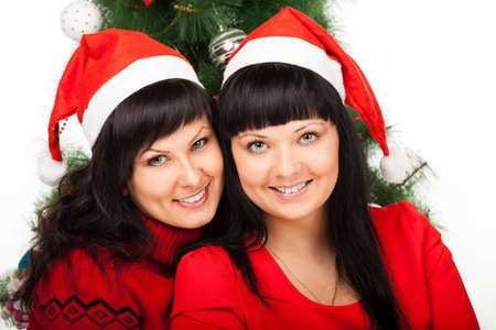 Two young dark-haired women in Santa s red caps smile near Christmas treeの写真素材