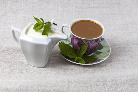 Cup of coffee on a saucer and a jar of cream, decorated with mint leaves on a background of gray fabric from flax.の写真素材