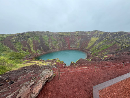 panoramic view of Kerid volcanic crater with turquoise crater lake and red rock mossy slopes with tiny figures on the hiking path on the rimの写真素材