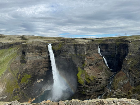panoramic view of Haifoss and Grannifoss plummet off a flat treeless plateau into a canyon showing different geological layers of stoneの写真素材