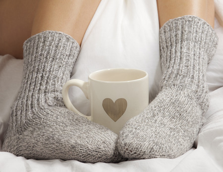 A cup of coffee or hot chocolate and female feet with socks on a white sheets