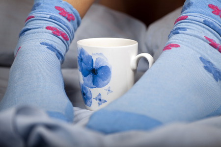 A cup of coffee or hot chocolate and female feet with socks on a blue sheets.