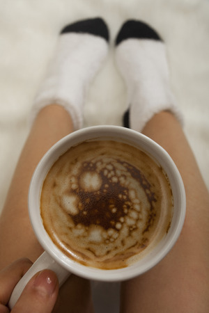 A cup of coffee or hot chocolate and female feet with socks on a white fur texture.の写真素材