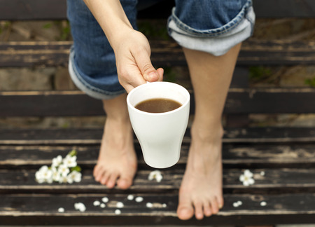Girl with bare feet drinking coffee or hot chocolate on a brown bench with white blossoms. The focus is on the cup.の写真素材