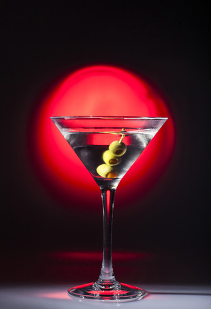 Sweaty glass of martini cocktail with olives on black background with red spot light.の写真素材