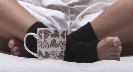 A cup of coffee or hot chocolate and female feet with socks on a white sheets.の写真素材