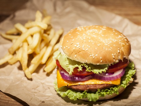Fresh delicious burger with cheese, tomato, onion, lettuce and french fries on brown paper on wooden tableの写真素材