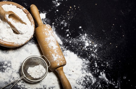 Metal strainer, rolling pin and wooden bowl with spilling flour on a black background.の写真素材
