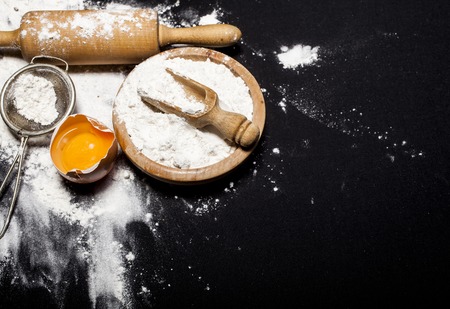 Ingredients and utensils for the preparation of bakery products - flour, dough, eggs, rolling pin, whisk, strainer, bread - on black table with free copy spaceの写真素材
