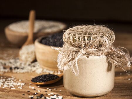 Fresh homemade sesame tahini in a glass jar and white and black seeds in wooden bowls and spoons on wooden backgroundの写真素材