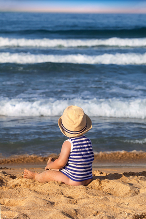 Baby boy with hat sitting on the beach near the water.の写真素材