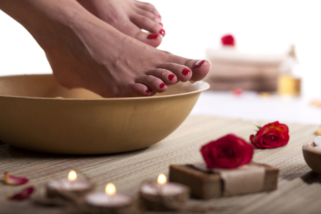 Female feet with drops of water, spa bowl, towels, flowers, candles, sea salt and bottle with essential oil on white background. Foot spa concept.の写真素材