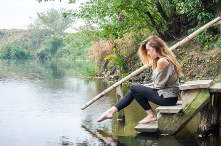 Young beautiful woman model with long blond hair sitting with different emotions laughter, sadness, sorrow, thoughtfulness on a wooden bridge on the bank of the river and the forest looking for waterの写真素材