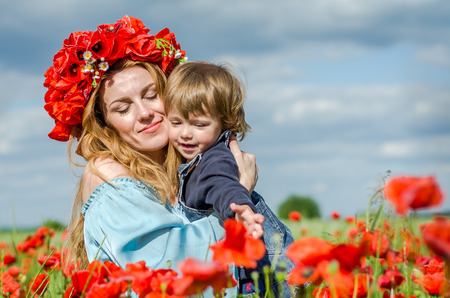 Young charming woman with long hair mom with her daughter in her arms walking in poppy field with a bouquet of poppies wreath on his headの写真素材