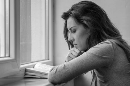 Young charming girl student, with long hair, sad at the window reading book with lessonsの写真素材