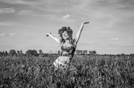 A young, happy, beautiful girl is having fun and dancing with joy in the field of flowering poppies with a wreath of poppy flowers on her head, a hot, sunny summer dayの写真素材
