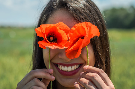 Young charming brunette girl in a field with flowers of poppies and daisies on a bright sunny summer dayの写真素材