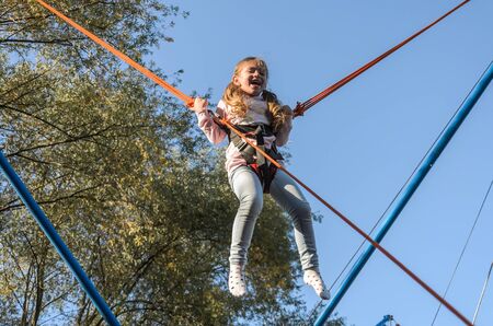 Little charming girl child on elastic ropes jumps on a trampoline in an amusement parkの写真素材