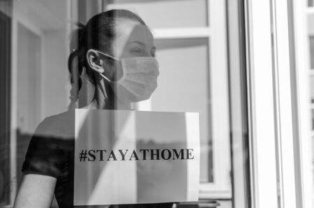 A young girl in a medical mask stands at the window with a sign saying stayathomeの写真素材