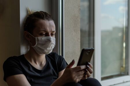 A young girl in a medical mask sits on a windowsill near a window and reads a bookの写真素材