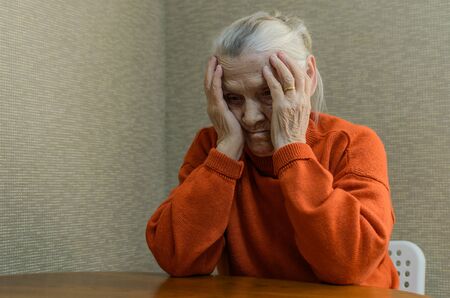 A lonely elderly woman sits at a table and holds her head sad from negative emotionsの写真素材