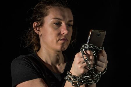 Young woman with chain tied hands to smartphone on black background