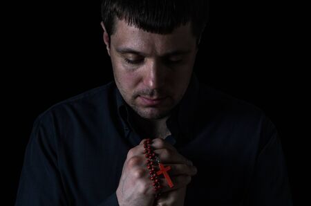 Portrait of a young man who praying with prayer beads with a crucifix on a cross on a black backgroundの写真素材